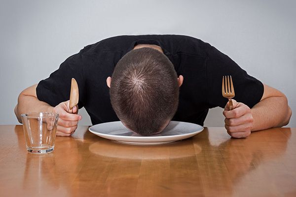 Hungry man, laying his head on an empty plate, holding a fork and knife in each hand.