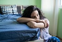 A woman leaning against her bed with her arms folding, looking deeply troubled.