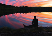 A man sitting at the edge of a lake, contemplating the sunset