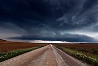 A long dirt road stretches to the horizon, where a large storm is brewing